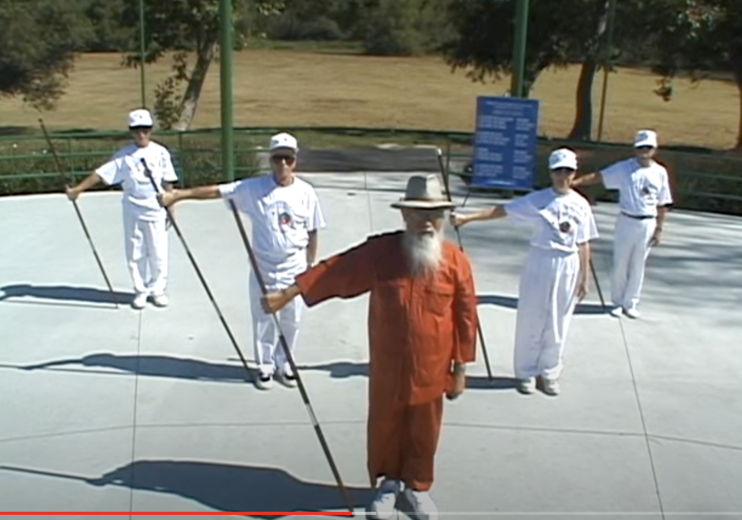Senior Vietnamese man with white hat, holding stick, teaches an exercise class outdoors
